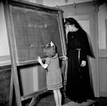 823245 Afbeelding van een Zuster Augustinesse, bij het schoolbord waarop een meisje een regel schrijft, in de ...